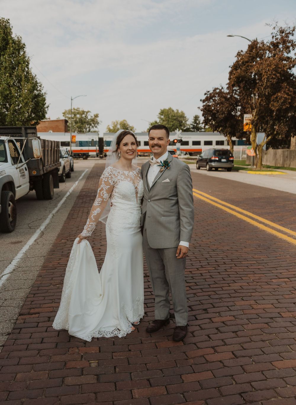 A couple in wedding attire poses on a brick paved street with a train visible in the background.