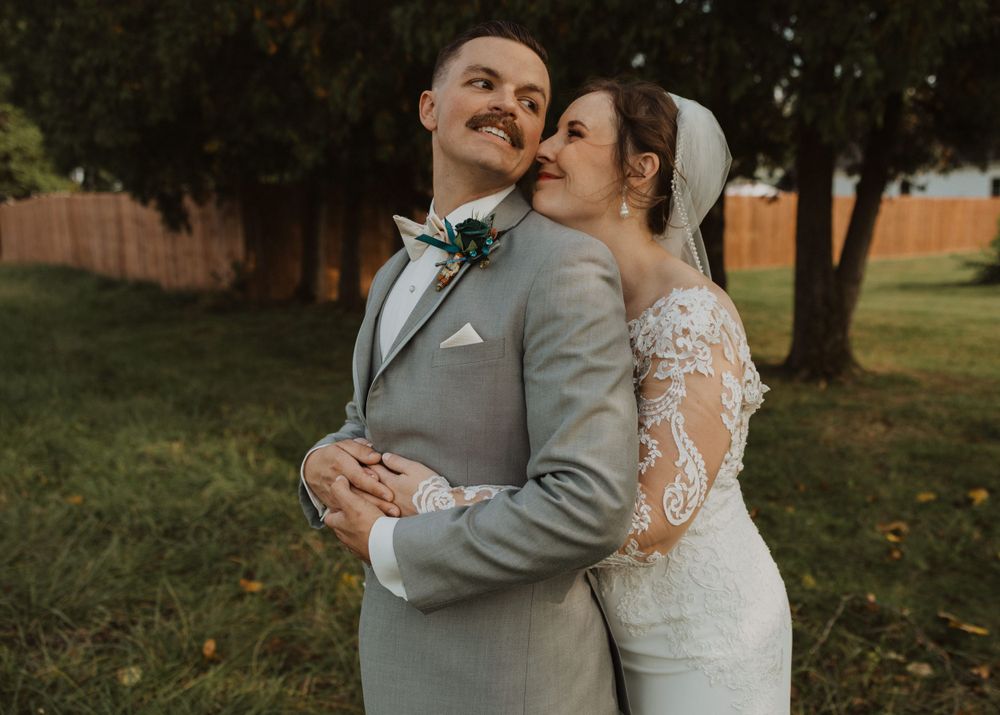 Wedding couple poses together on grassy lawn during sunset with trees creating a natural backdrop.