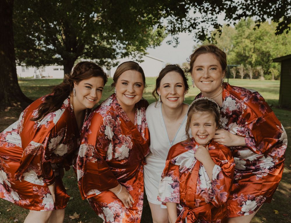 Wedding party wearing matching red floral robes pose together outdoors under a tree.