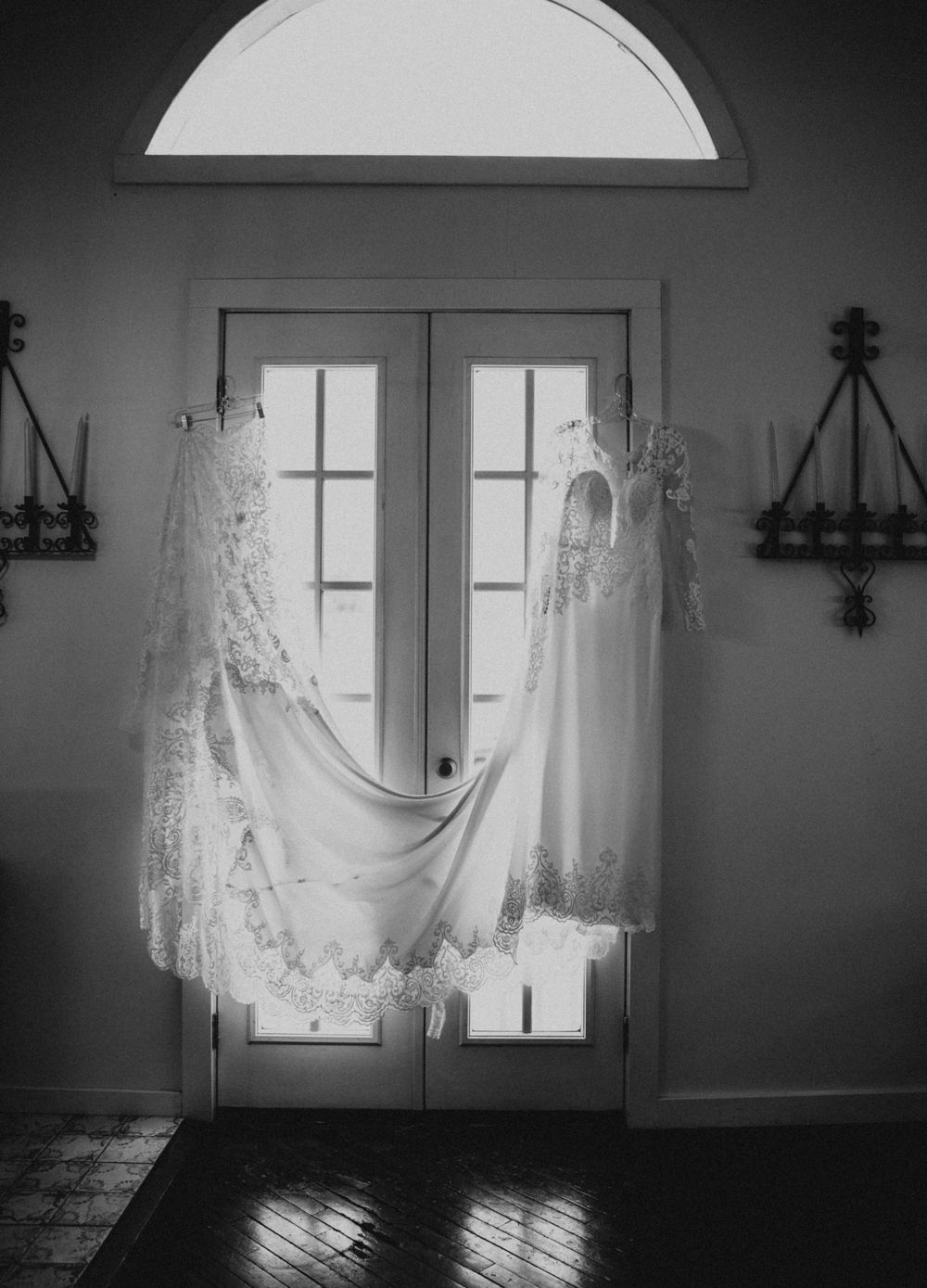 Two elegant white wedding dresses hanging in front of French doors with natural light streaming in.