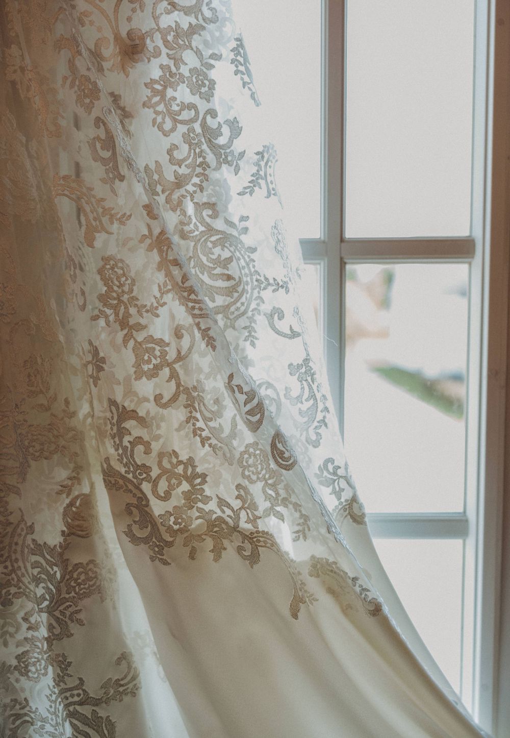 Close-up detail of intricate lace pattern on white wedding dress near a window.