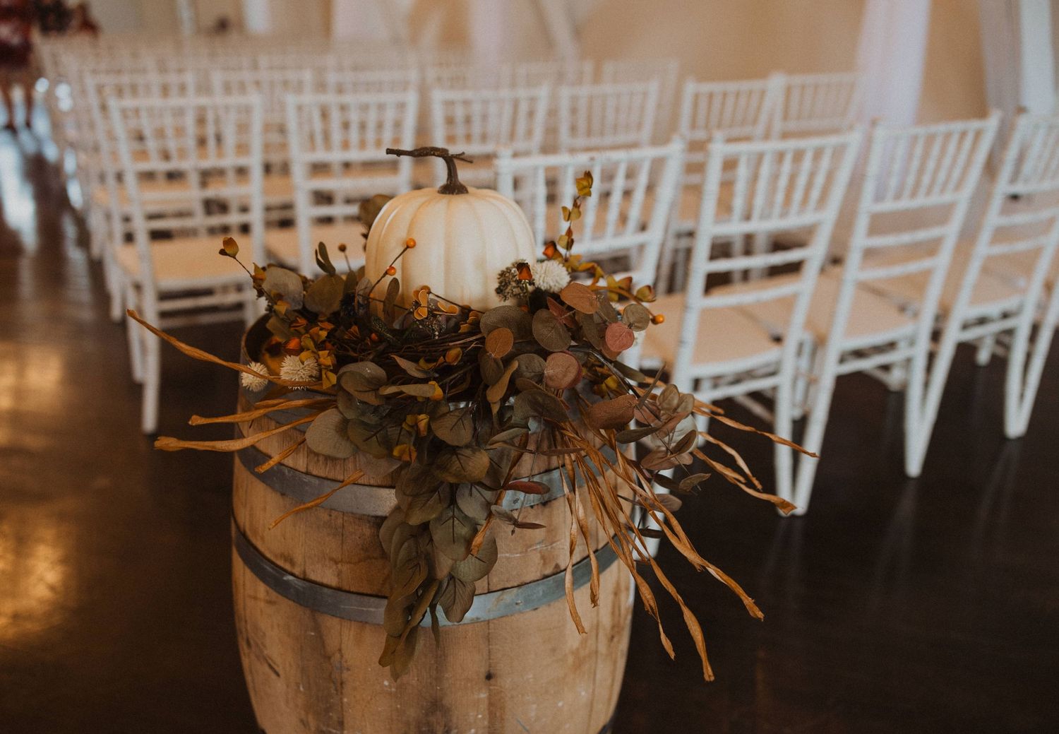 White pumpkin and floral arrangement on rustic barrel next to Chiavari chairs at wedding ceremony.