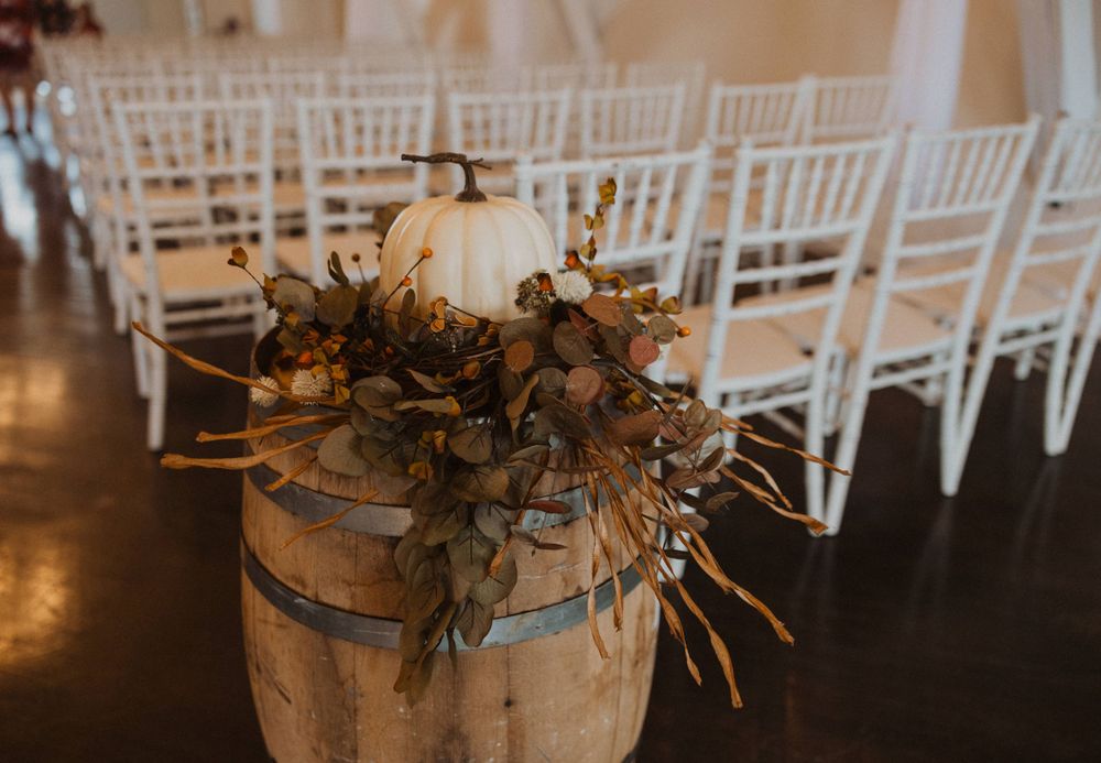 White pumpkin and floral arrangement on rustic barrel next to Chiavari chairs at wedding ceremony.