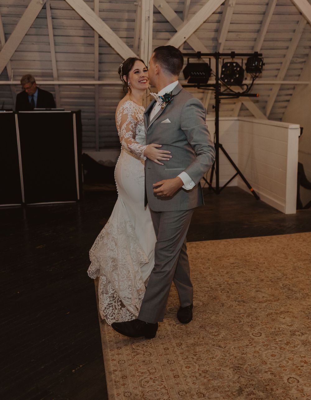 A couple shares a romantic dance moment in a rustic barn venue with wooden beams and warm lighting.