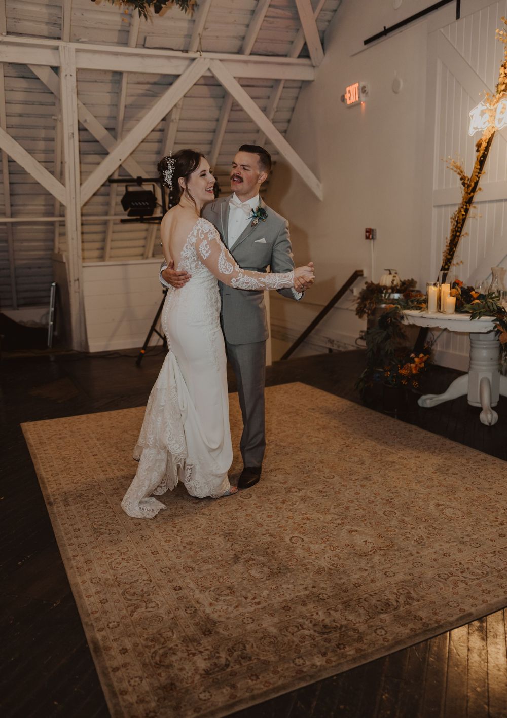 A wedding couple performs their first dance on a rustic wooden floor under vaulted barn ceilings.