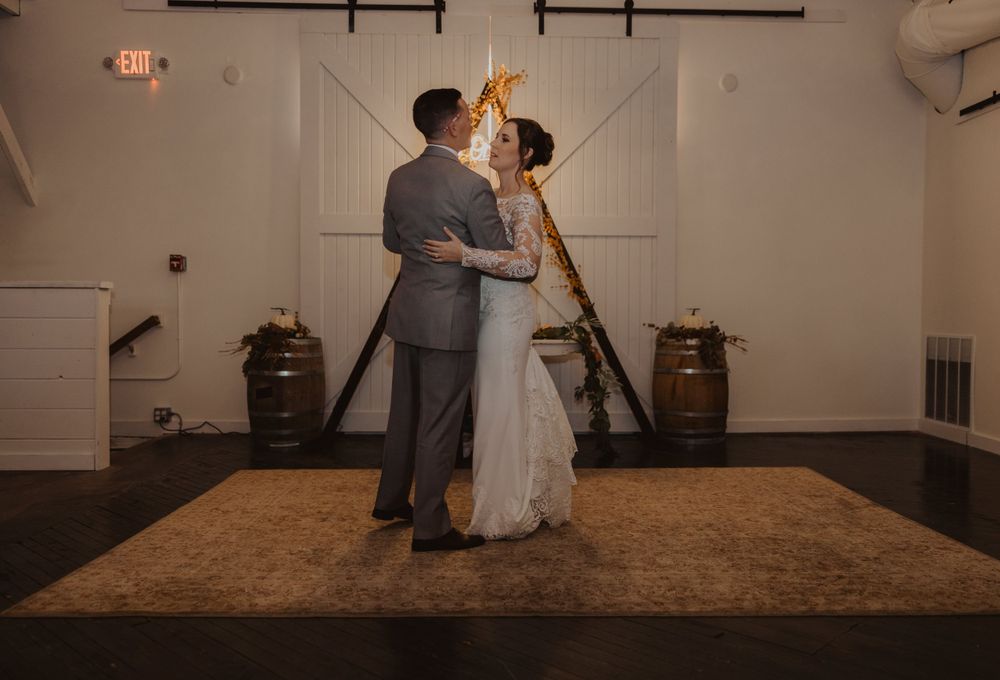 A couple dances beneath exposed beams in a warmly lit rustic wedding venue.