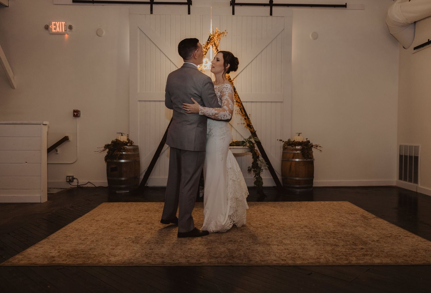 A couple dances beneath exposed beams in a warmly lit rustic wedding venue.