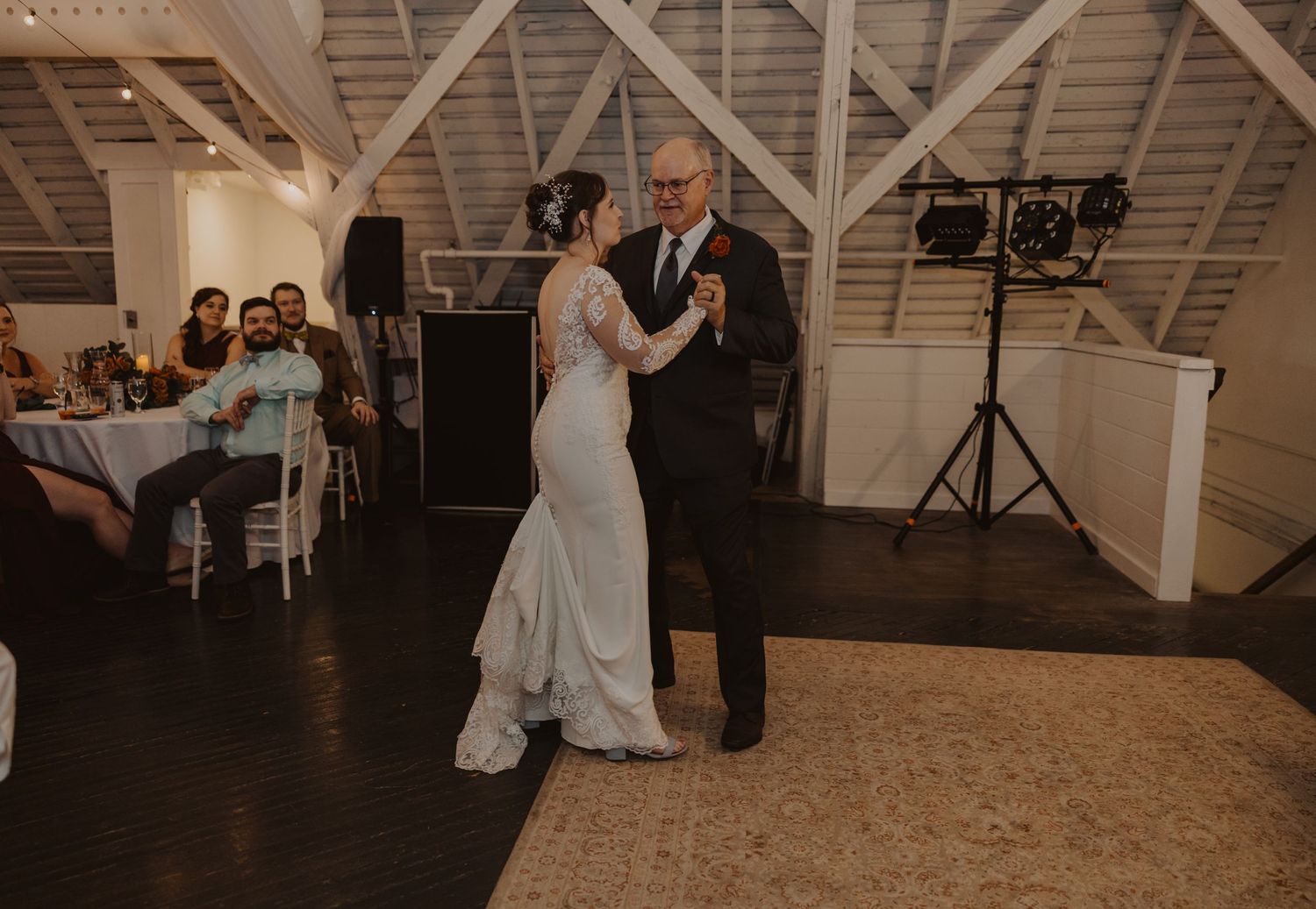 Wedding reception dance in a rustic barn venue with string lights and wooden ceiling beams.