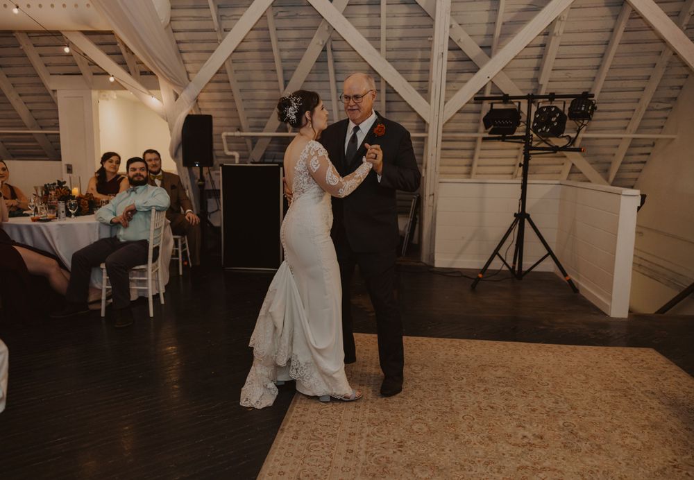 Wedding reception dance in a rustic barn venue with string lights and wooden ceiling beams.