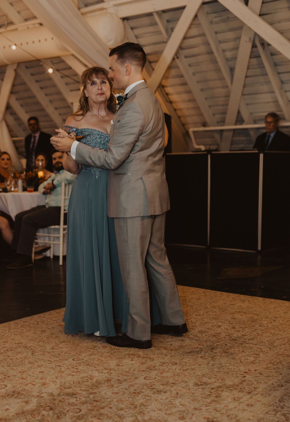 A couple shares a dance together at a wedding reception with warm lighting and exposed wooden beams overhead.
