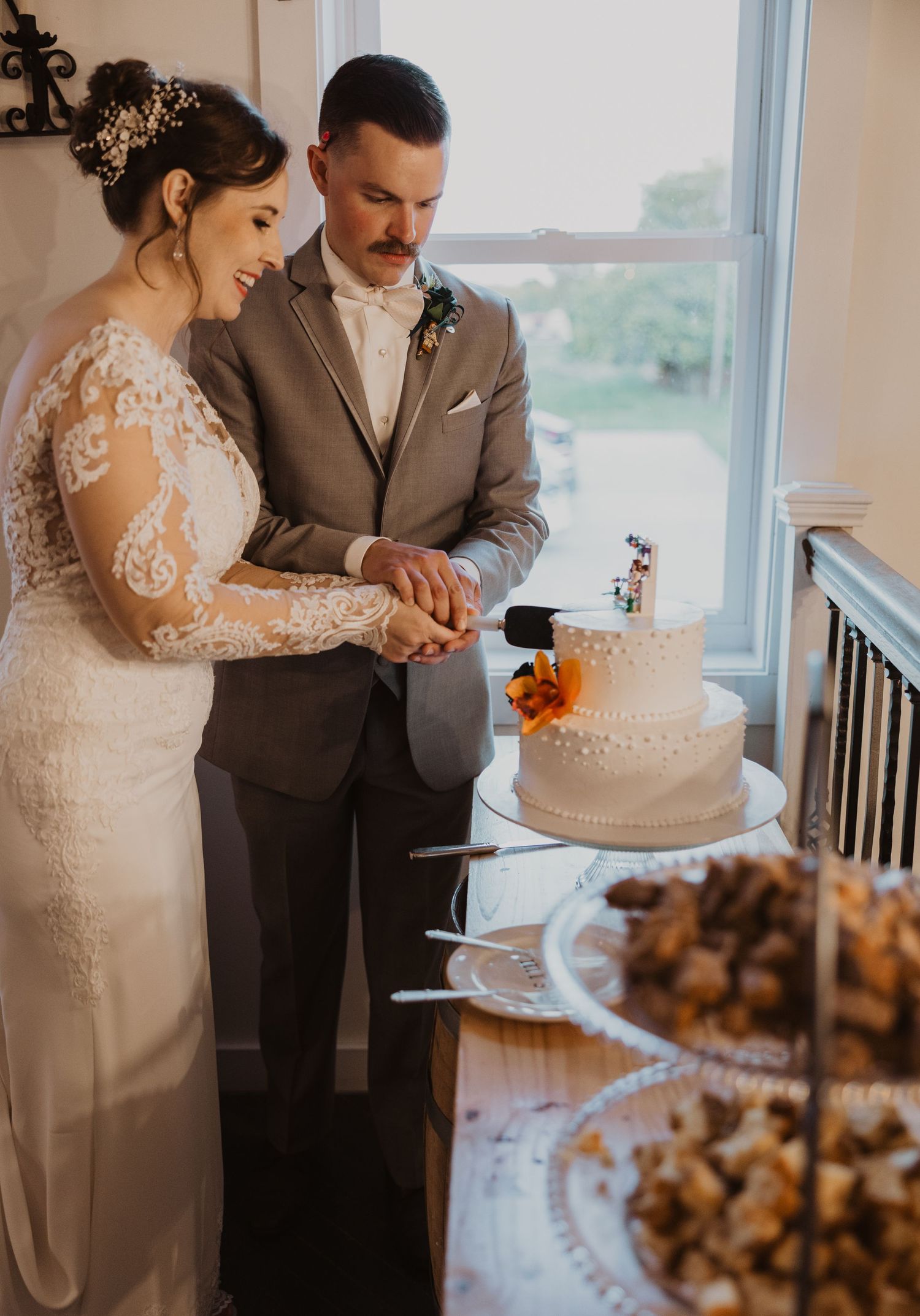 Newlyweds cut their white wedding cake together at a reception near a bright window.