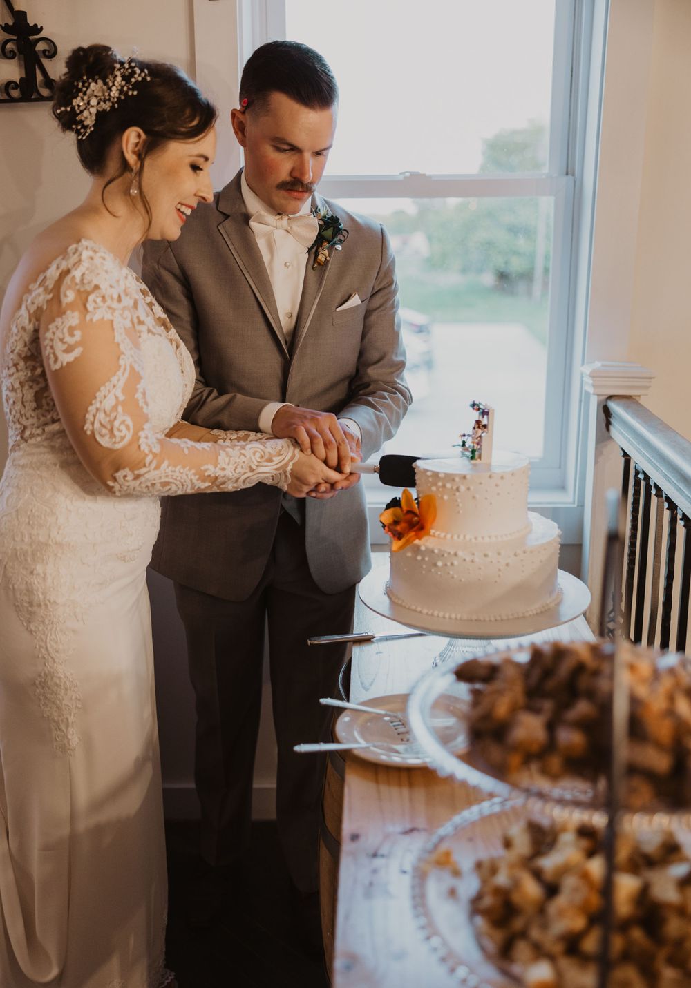 Newlyweds cut their white wedding cake together at a reception near a bright window.
