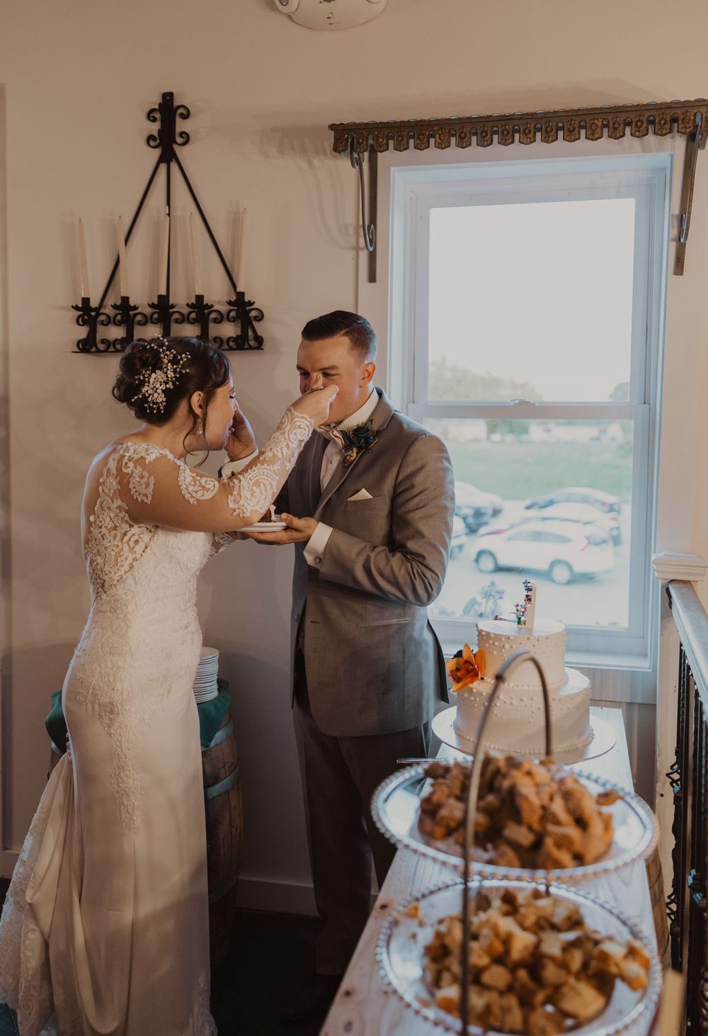 Couple sharing their first bite of wedding cake during their reception by a sunlit window.