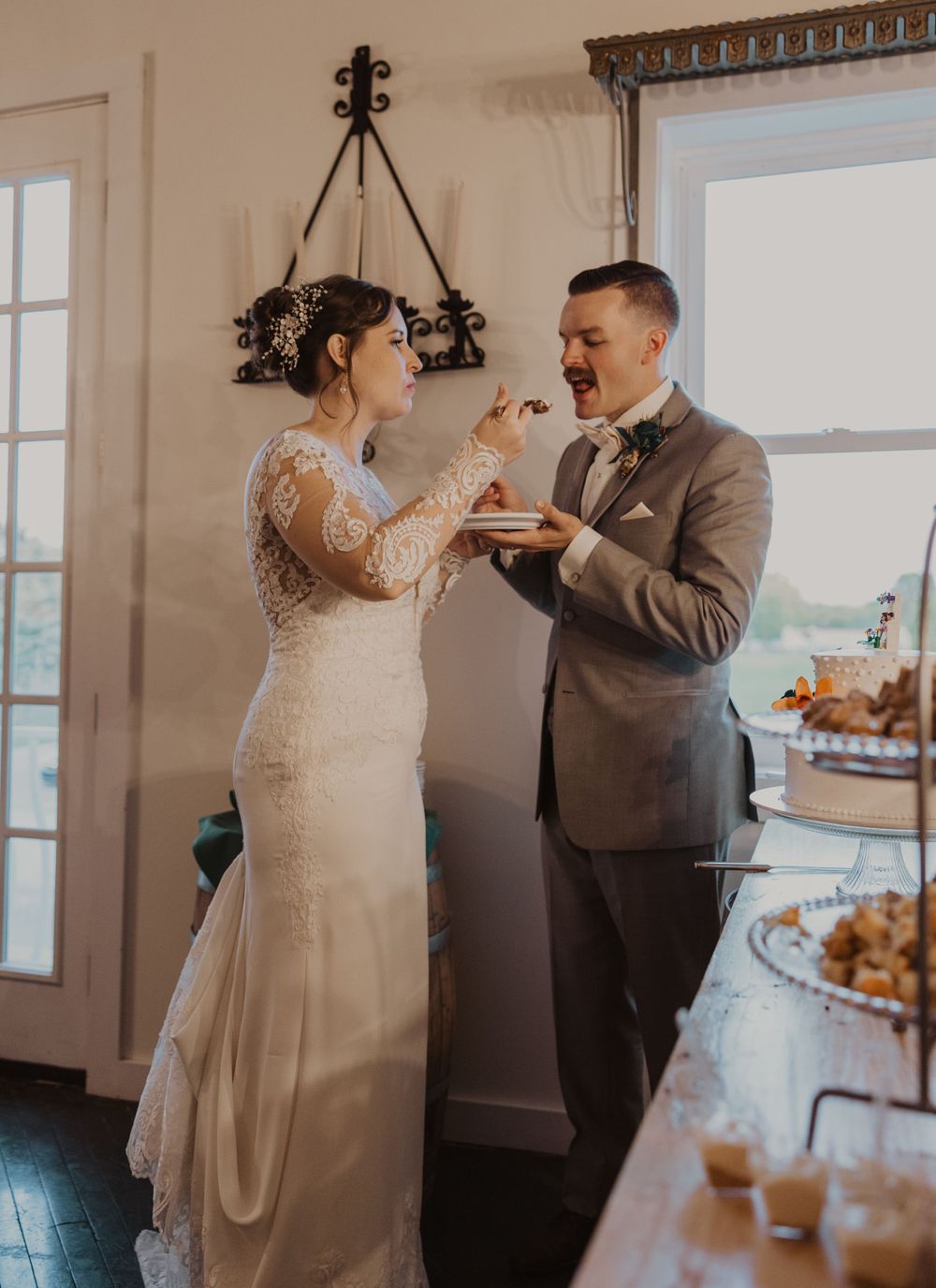 Newlyweds share cake during their wedding reception in front of a bright window.