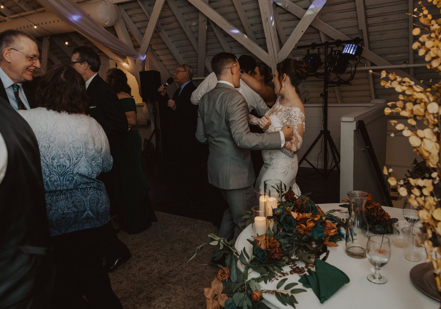 Wedding party dancing and mingling in a barn venue decorated with warm lighting and rustic decor.