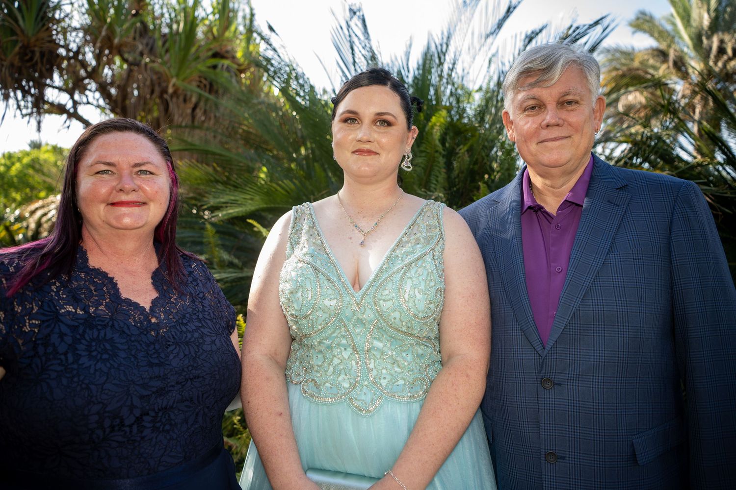 Three people pose together outdoors with palm trees visible in the background on a sunny day.