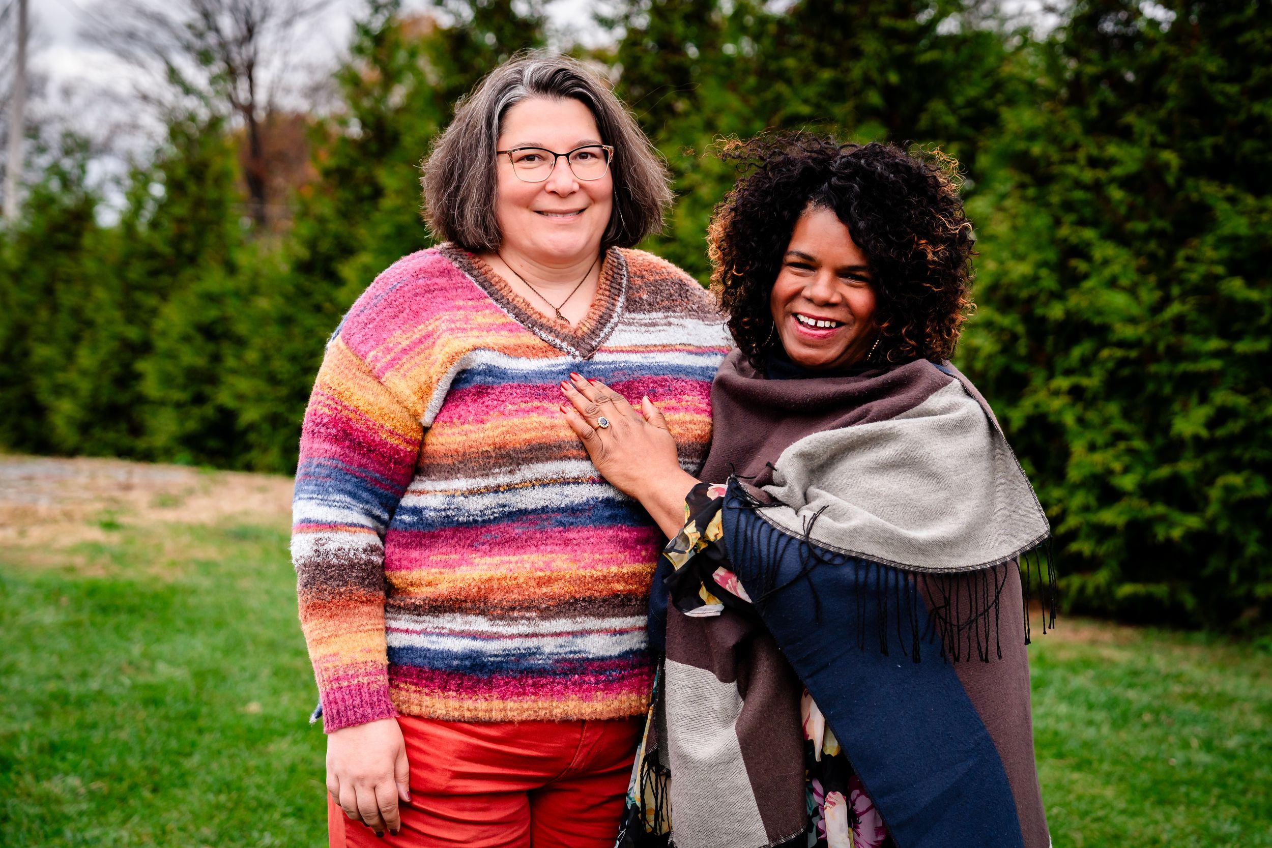 Two people stand together outdoors on a grassy hill with trees in the background wearing colorful sweaters.