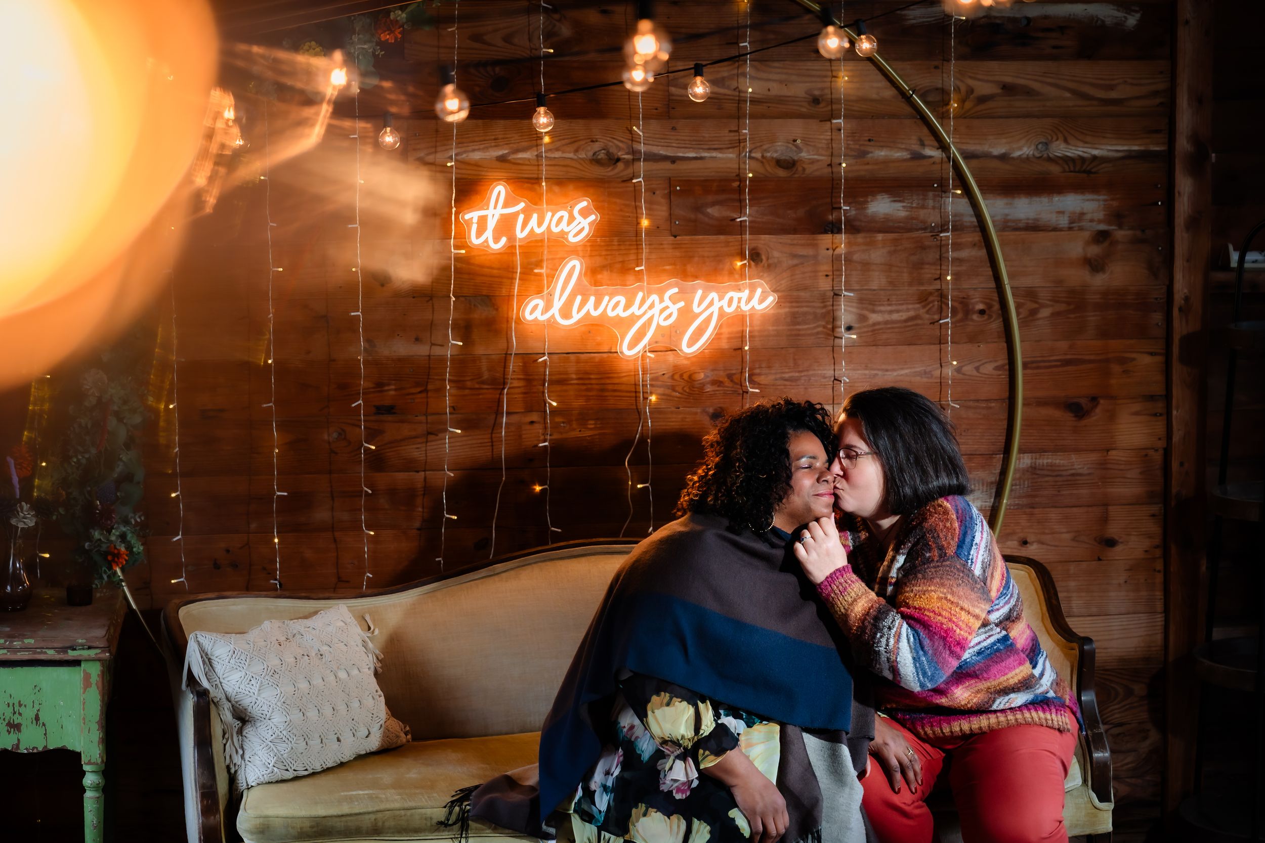 Couple embraces in a romantic moment under warm glowing lights against rustic wooden walls at an intimate venue.
