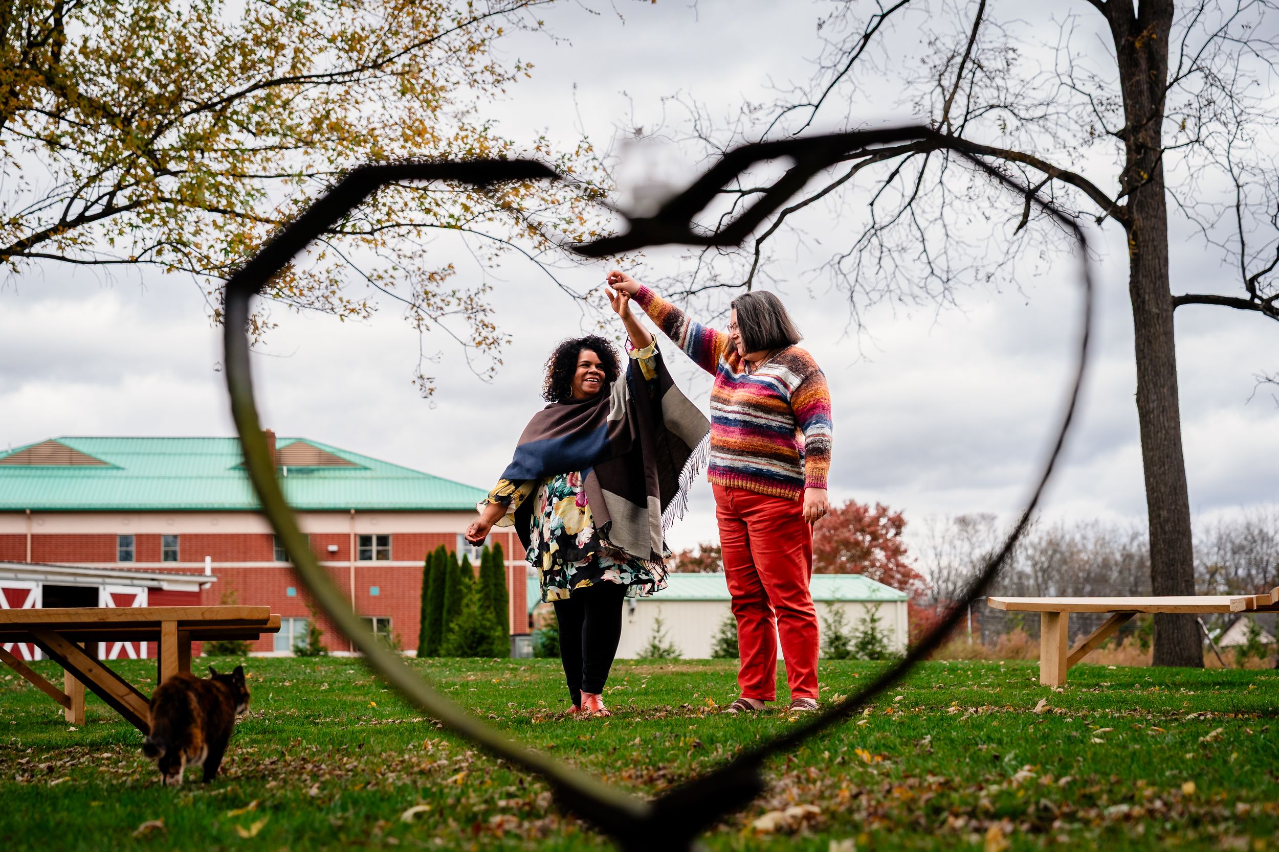 A creative photo through a heart-shaped frame shows two figures standing on grass with autumn trees in background.