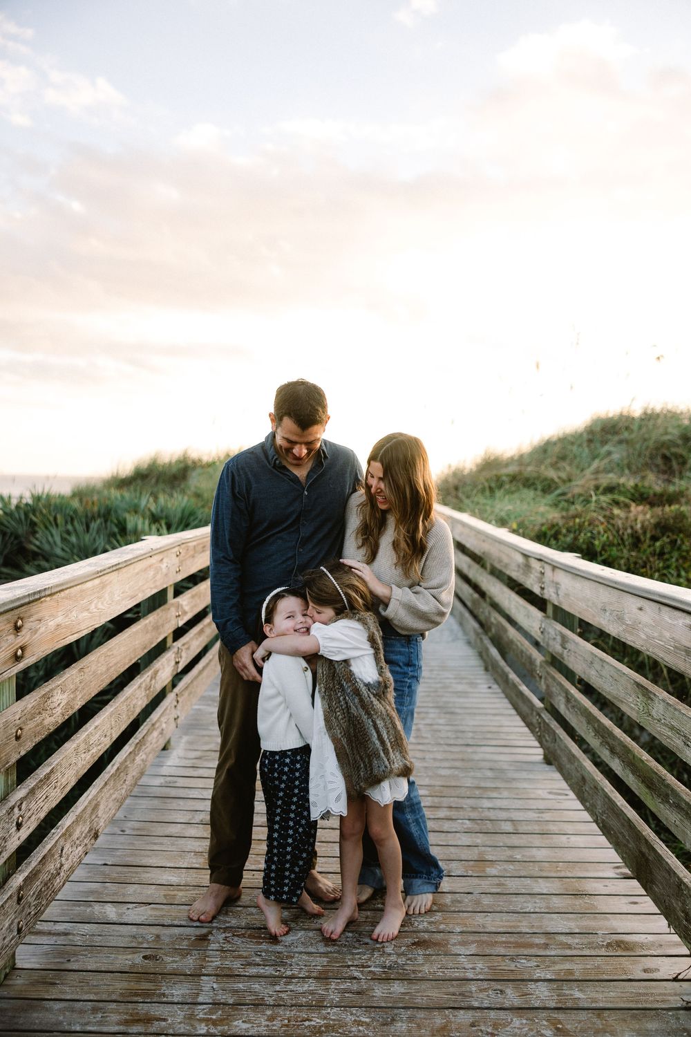 A family shares tender moments together on a wooden boardwalk during a sunset photoshoot at the beach.