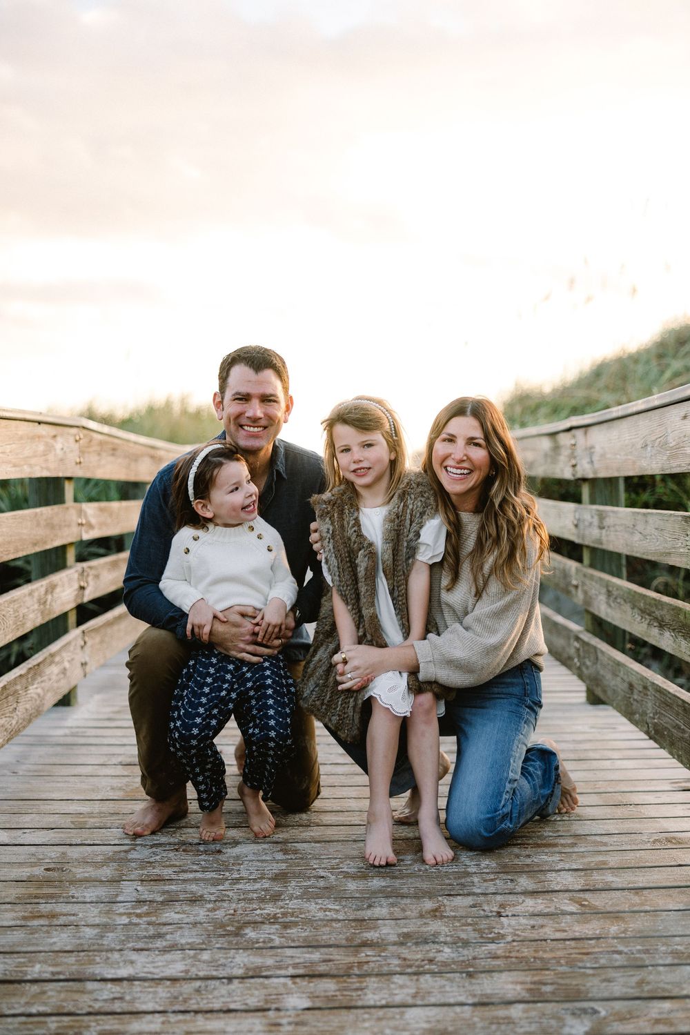 A family poses together on a wooden boardwalk during a casual outdoor photo session at sunset.