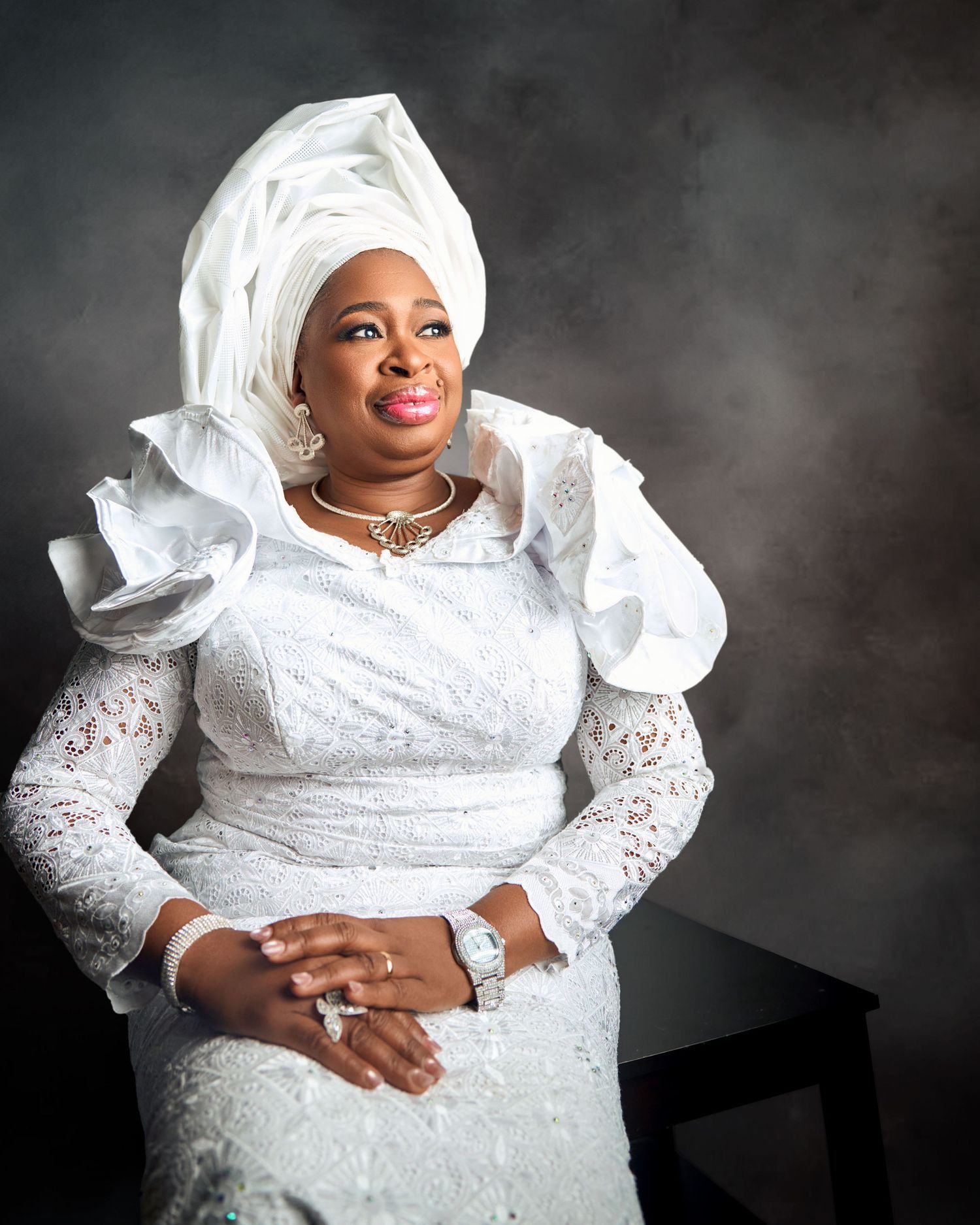 Woman in white Nigerian lace dress with white gele headwrap seated elegantly in a studio.