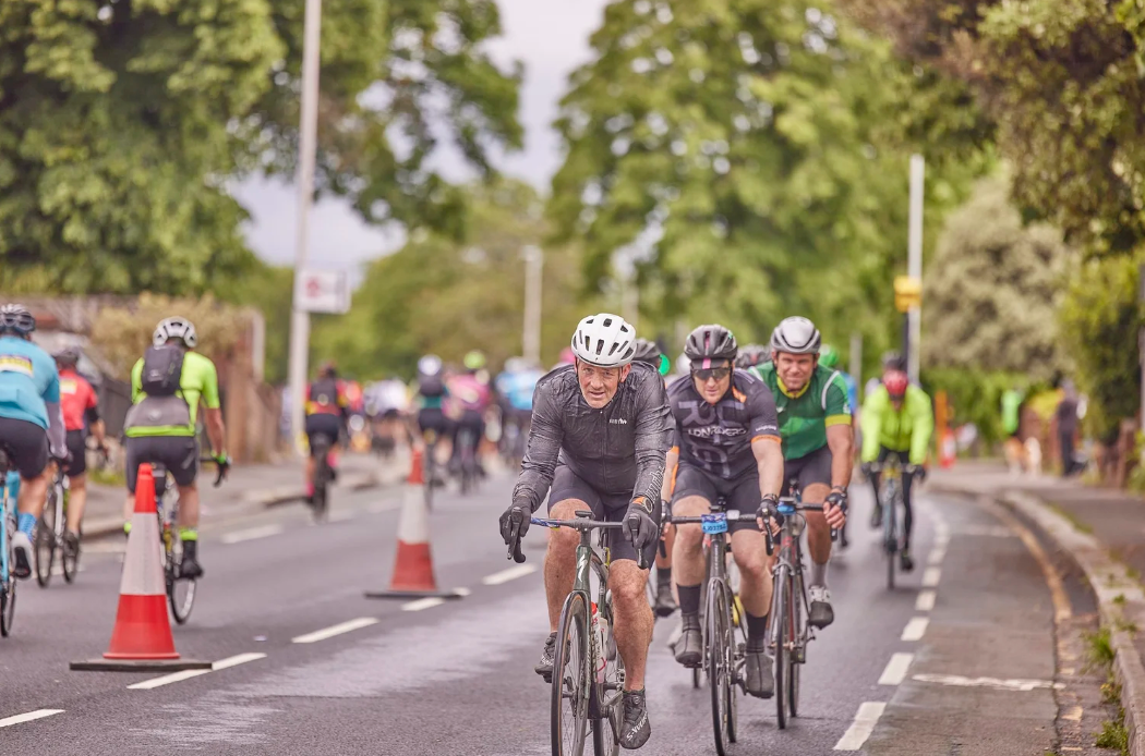 Cyclists compete in a road race along a tree-lined street with orange traffic cones marking the course.