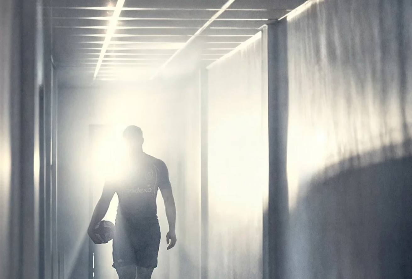 A silhouetted figure walks through a bright, misty corridor with glass walls creating a modern architectural atmosphere.