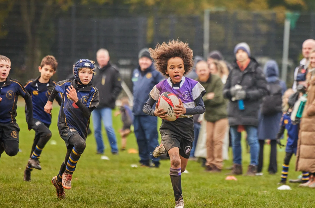 Rugby players sprint across the field during a competitive match with spectators watching.