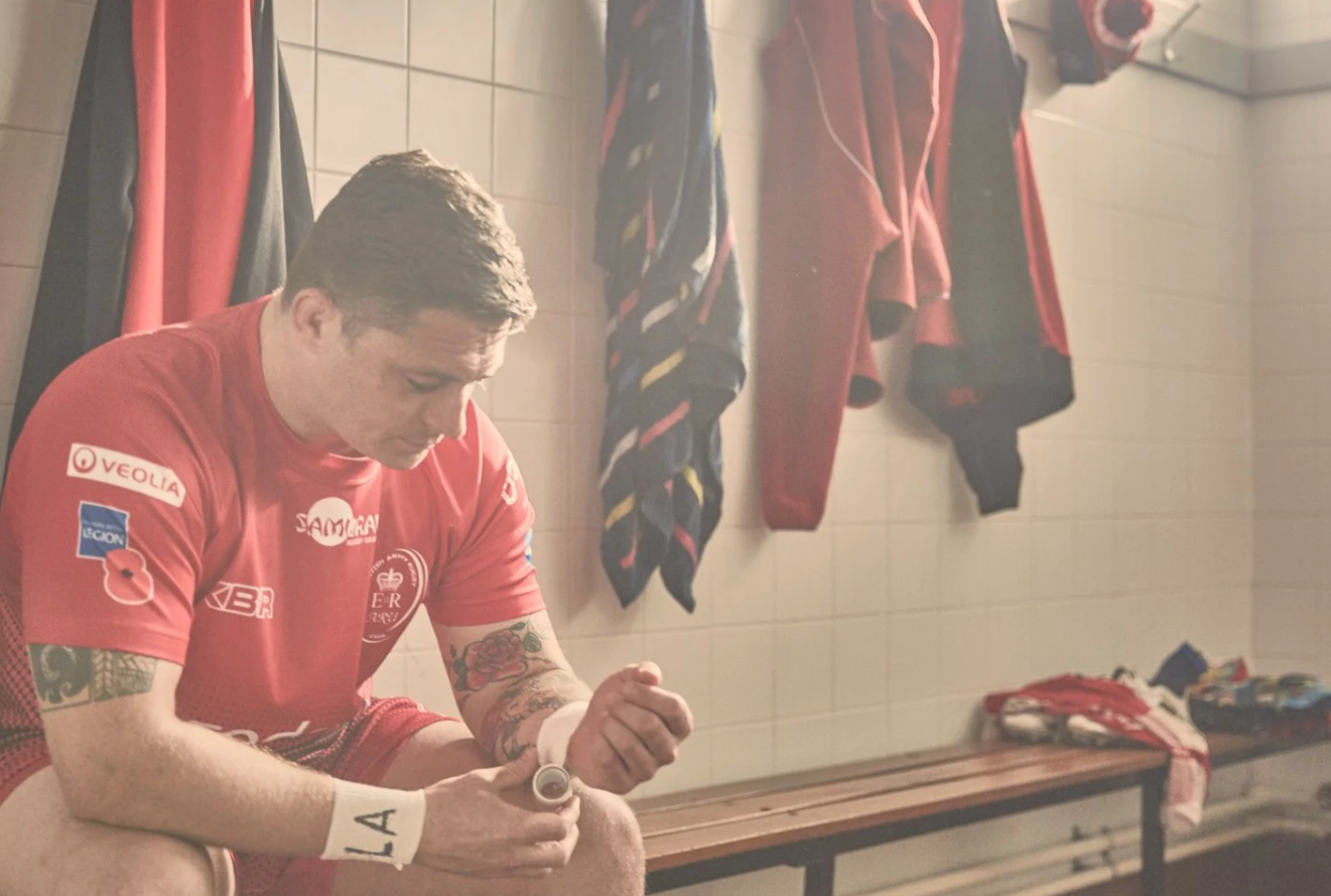 Soccer players in red jerseys sit in a locker room preparing for a match with sports equipment visible.