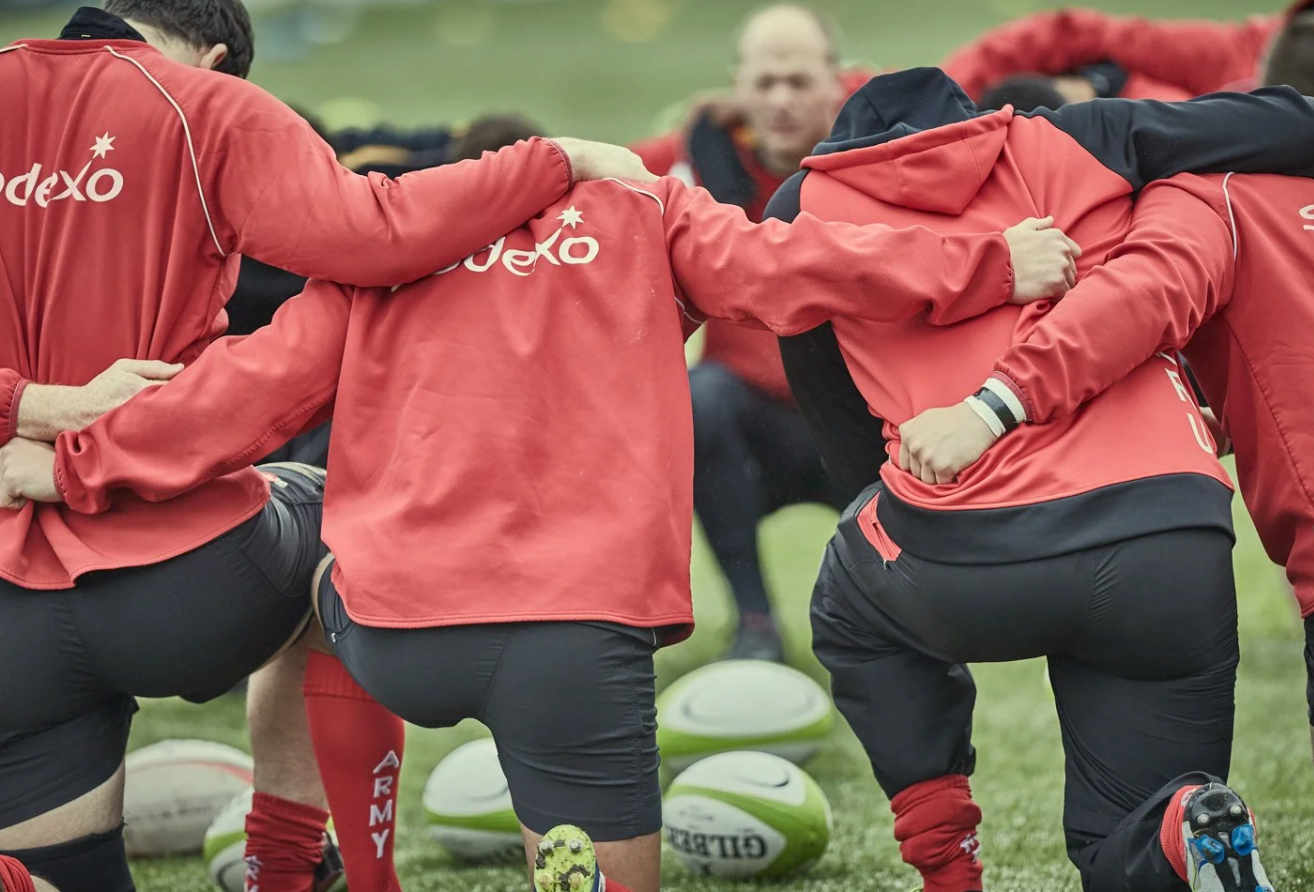 Rugby players in red jerseys perform stretching exercises during training session on green field.