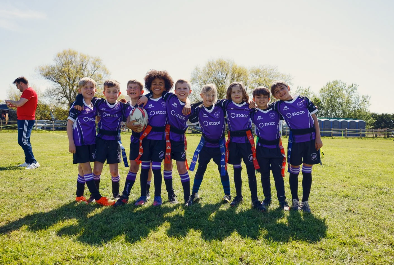 Youth soccer team players in purple jerseys line up together on a sunny grass field during practice.