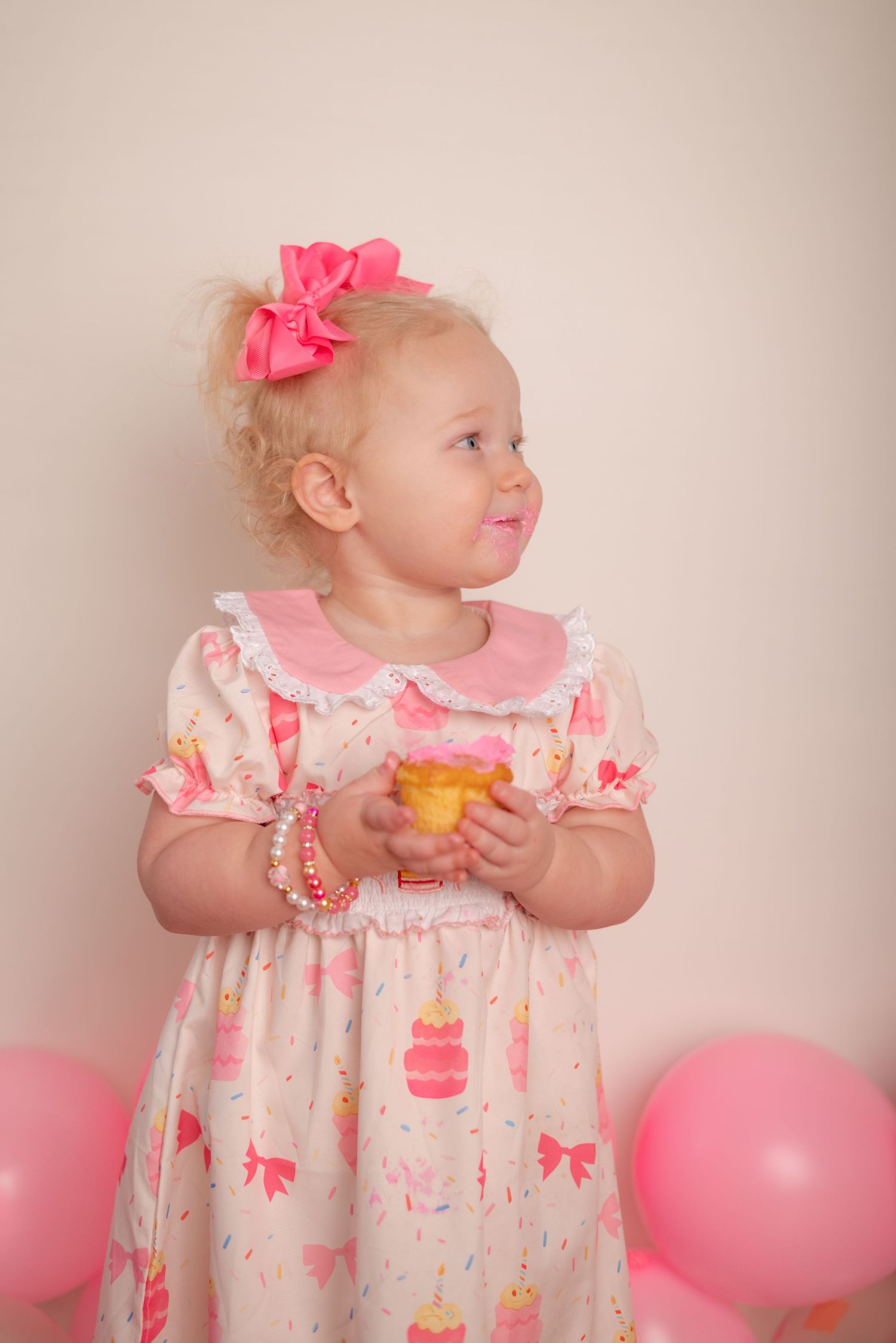 Baby in floral dress with pink bow holding orange snack while pink balloons decorate background.