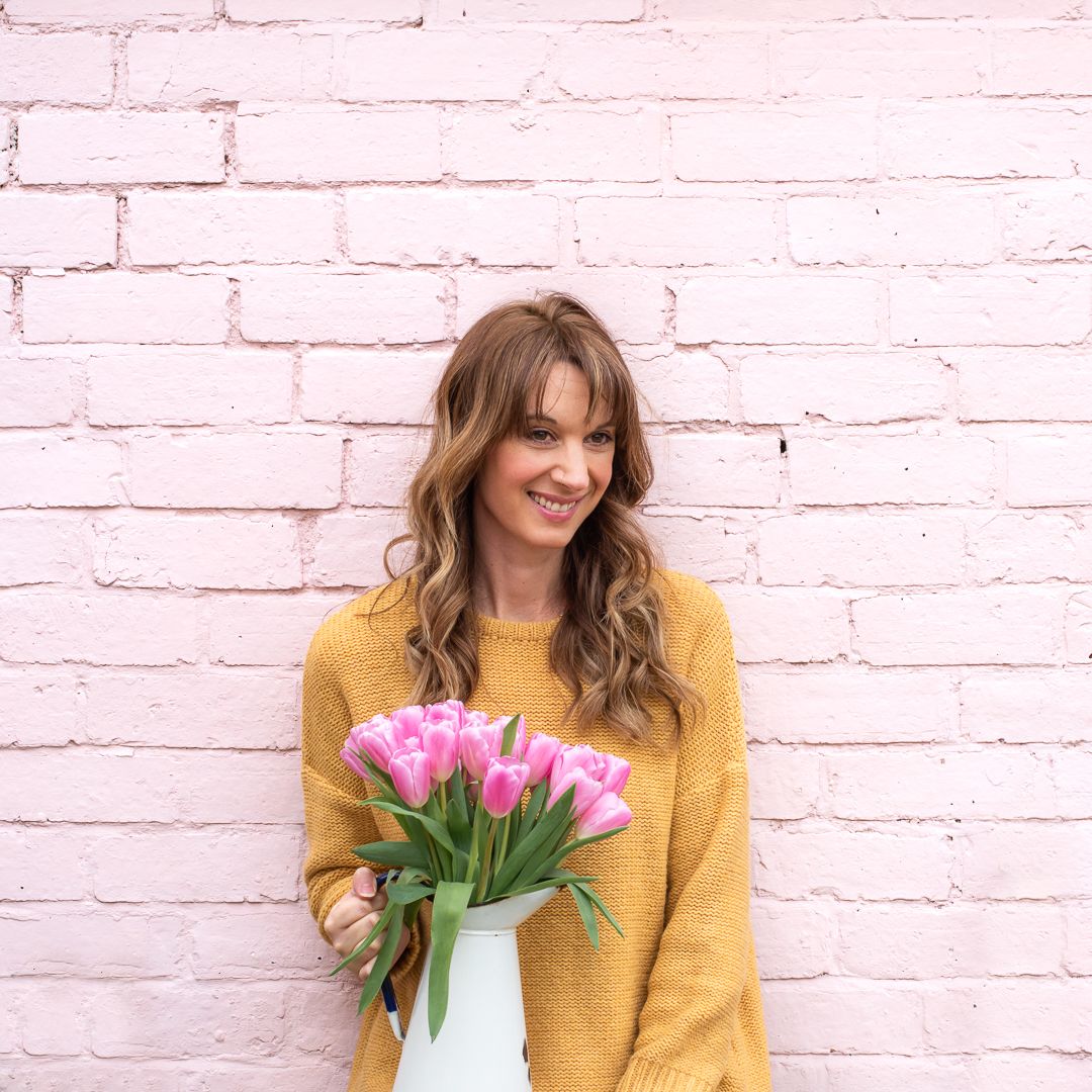 Woman (Hannah) smiling and holding a bouquet of flowers in front of a pastel pink wall.
