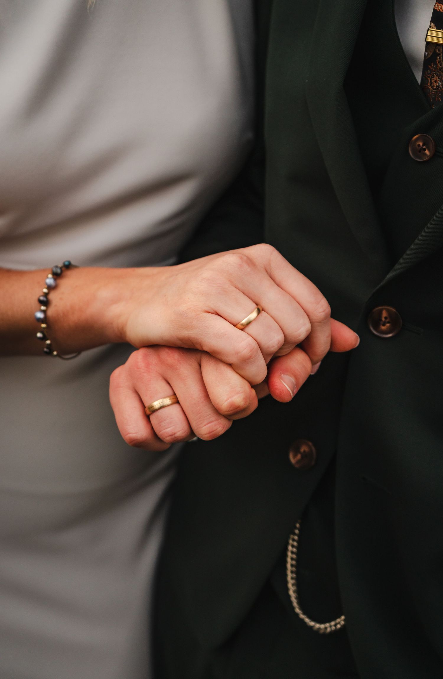 Close-up shot of two hands holding each other, with wedding rings and bracelets visible against a dark background.