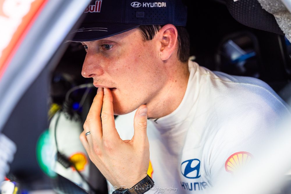 Driver in racing suit and cap sits in cockpit of race car with Hyundai branding visible.
