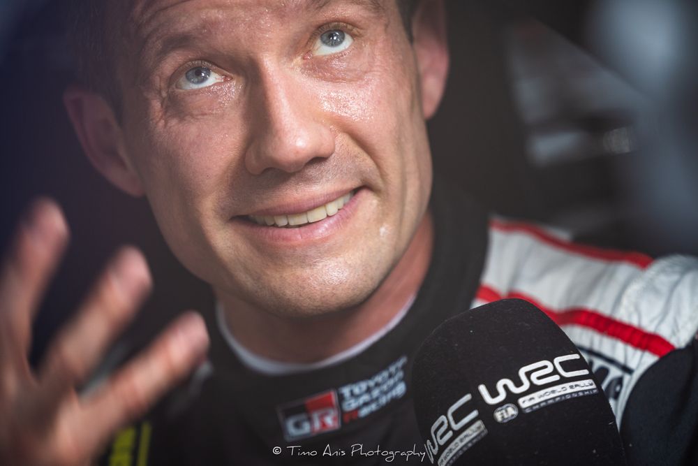 A close-up portrait of a smiling racing driver wearing a black and white racing suit.