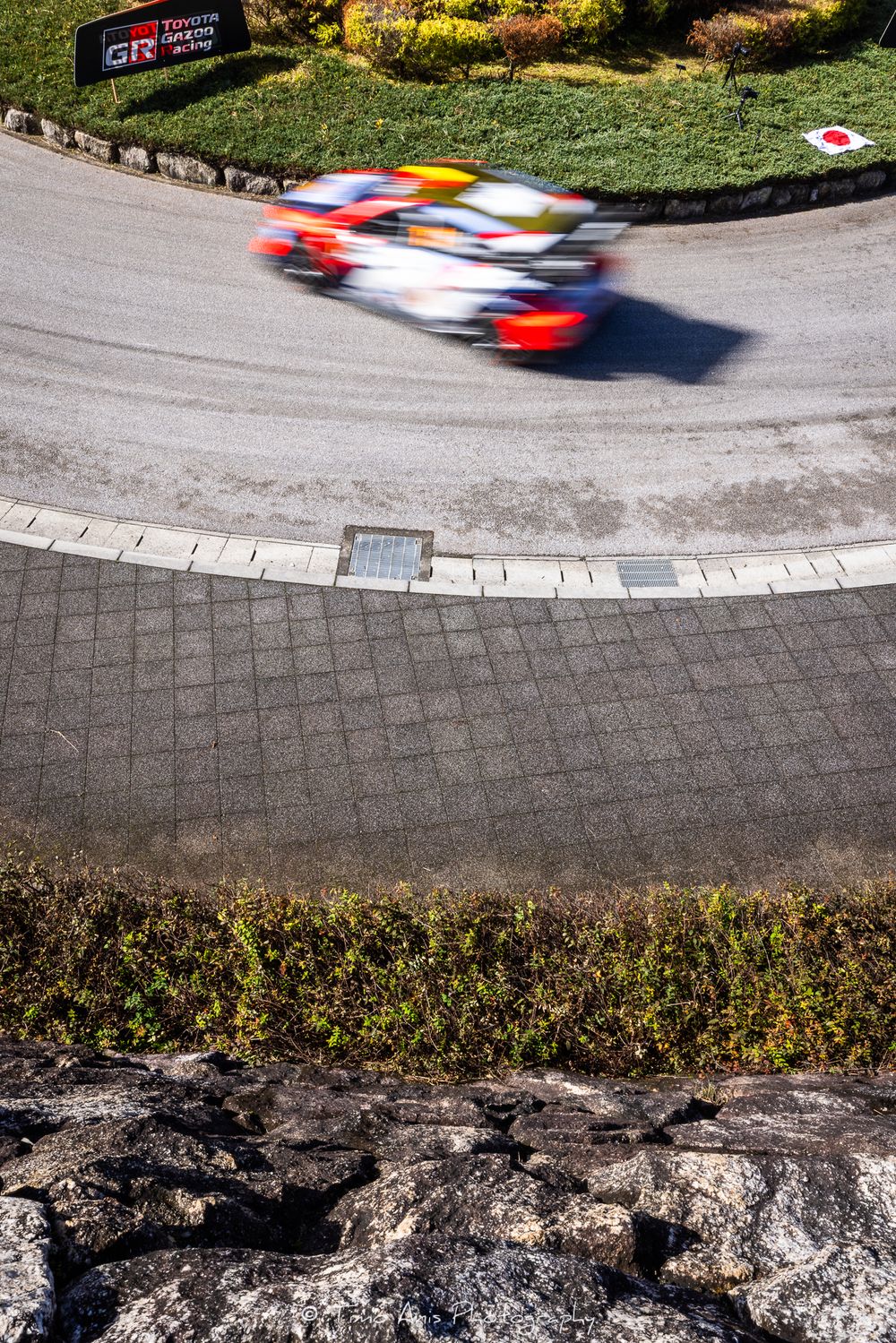 A race car speeds around a winding mountain road curve with motion blur against a rocky cliff backdrop.