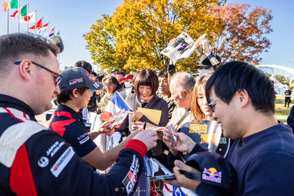 A crowd of people gathered outdoors during a fall day with colorful autumn trees in the background.
