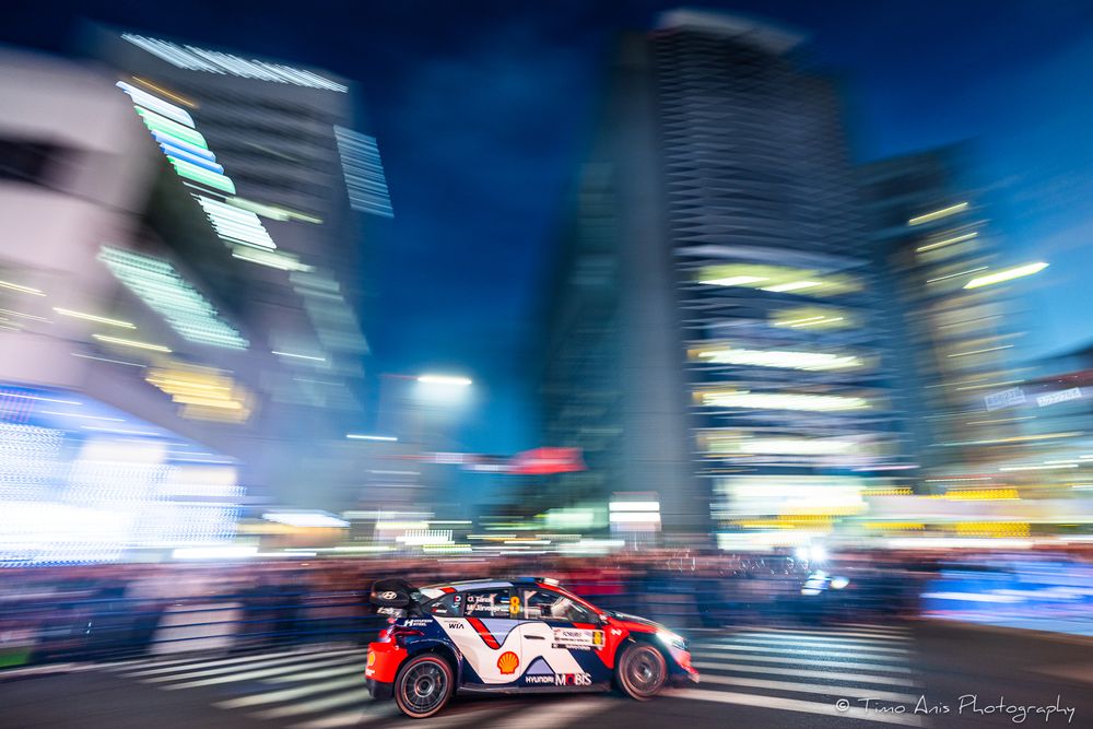 A rally car speeds through a blurred cityscape at night with illuminated skyscrapers in the background.