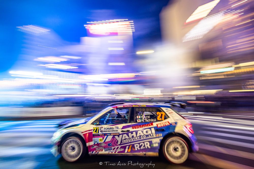 Rally car racing through city streets at night with motion blur and streaking lights creating dynamic movement effect.