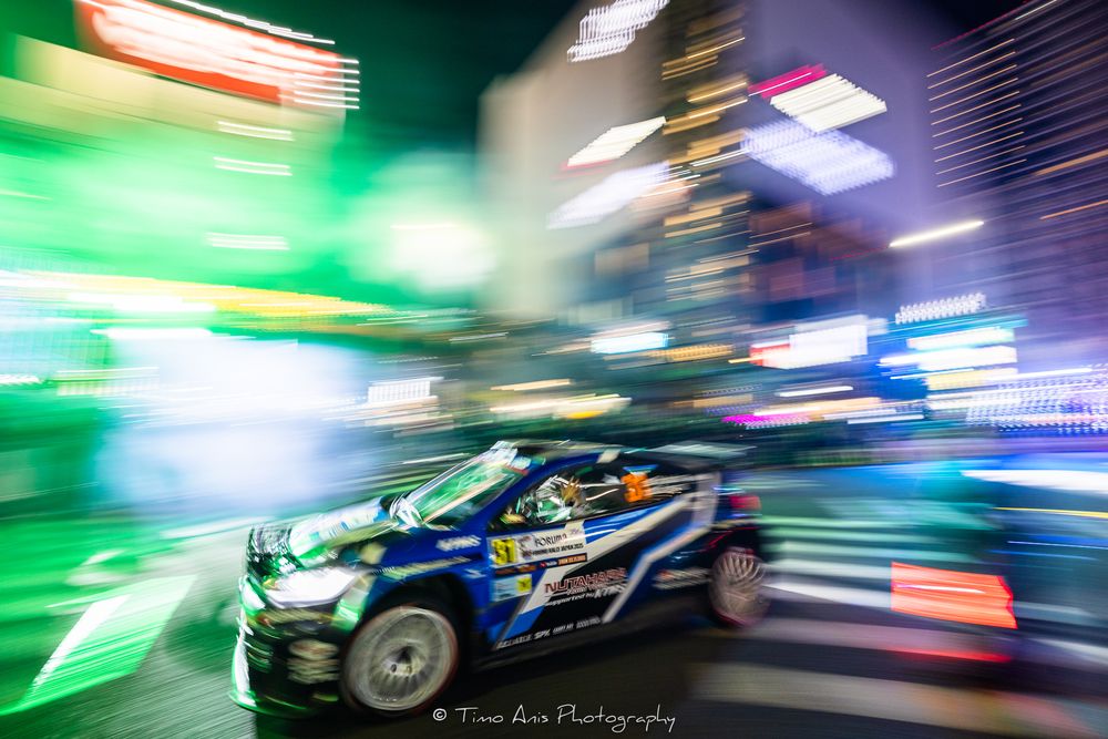 Motion blur racing scene with green and blue lights streaking across the frame during a nighttime motorsport event.