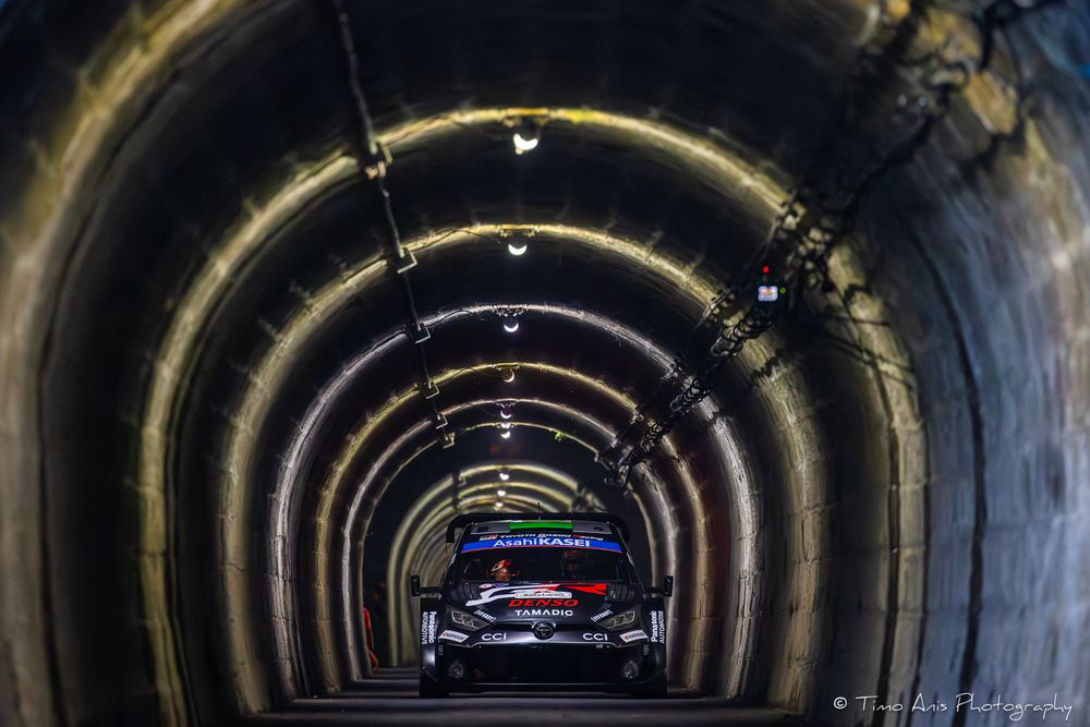 Rally cars racing through illuminated tunnel with curved stone walls and dramatic shadows.