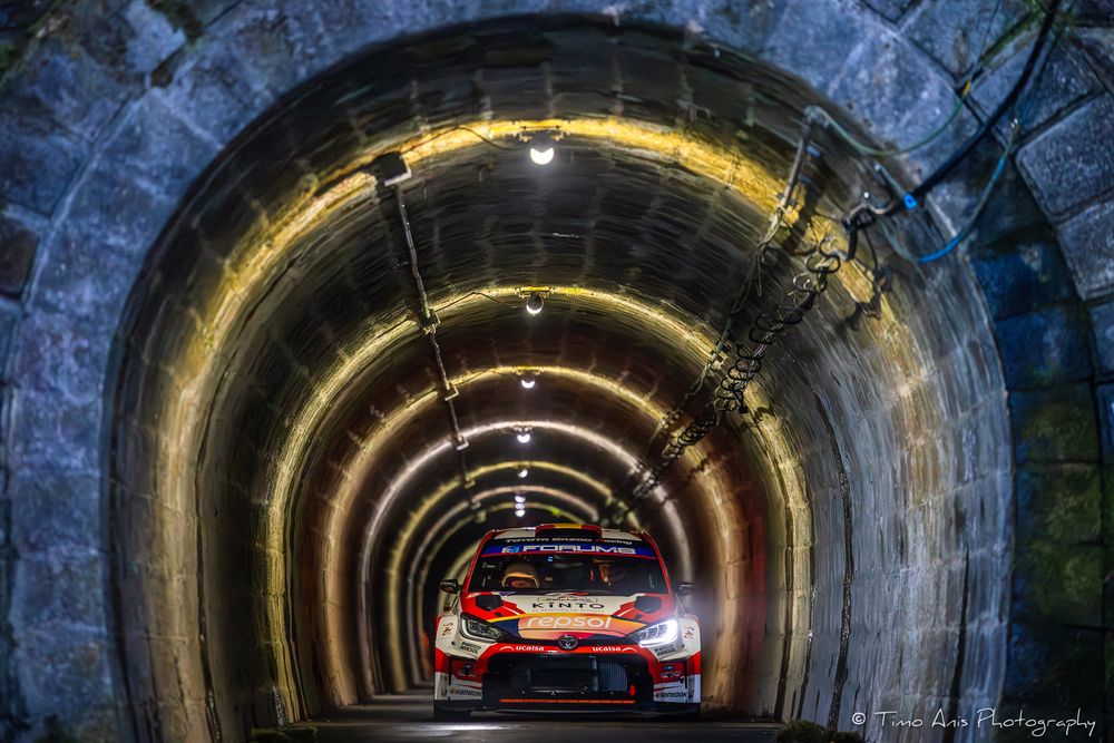 Red rally car emerges from dark stone tunnel during motorsport competition with dramatic lighting effect.