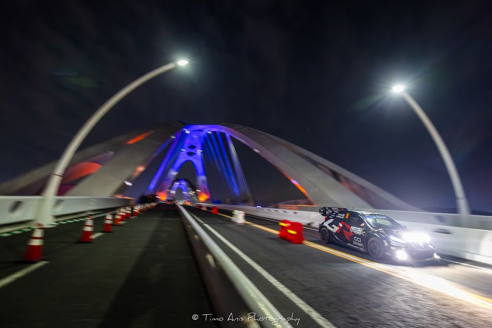 Illuminated highway bridge at night with red taillights creating light trails and blue architectural lighting.