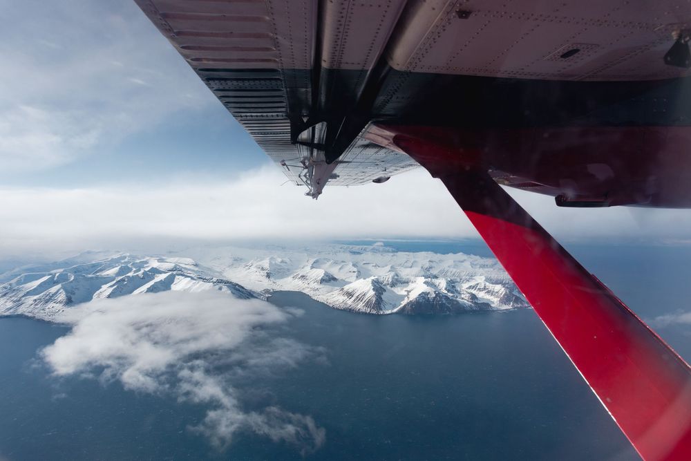 Aerial view from small aircraft showing red wing and snowy mountain peaks over ocean with scattered clouds.