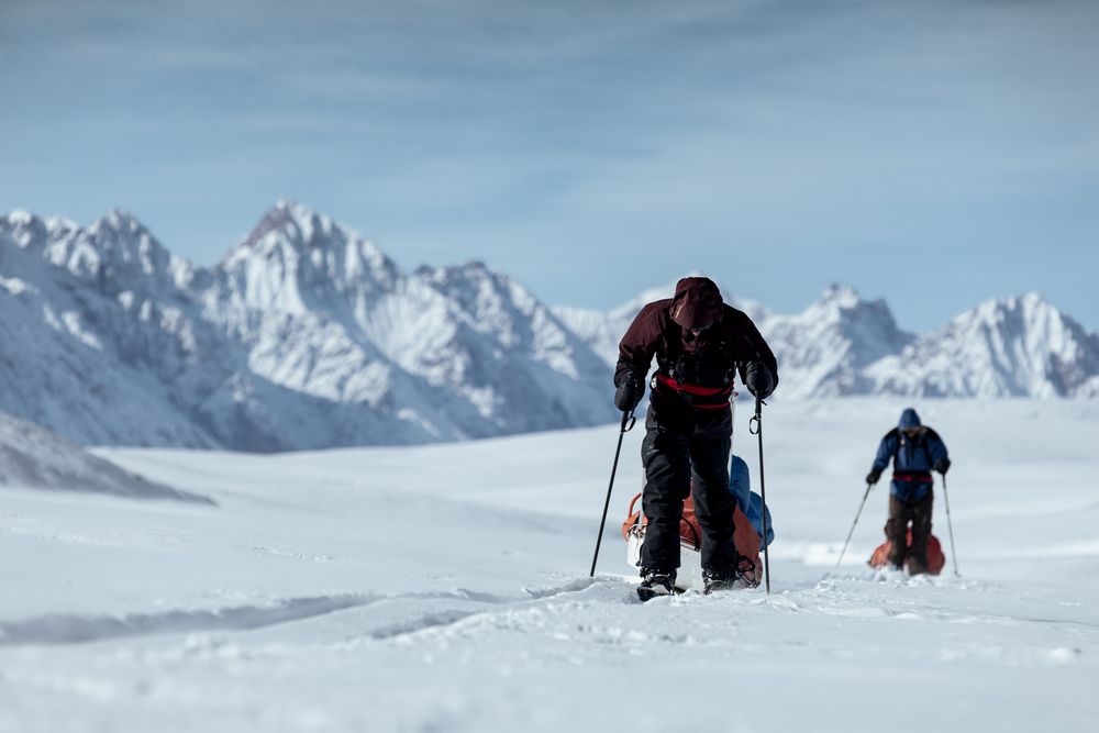Skiers traverse across a pristine snowy mountain slope with jagged peaks in the background.