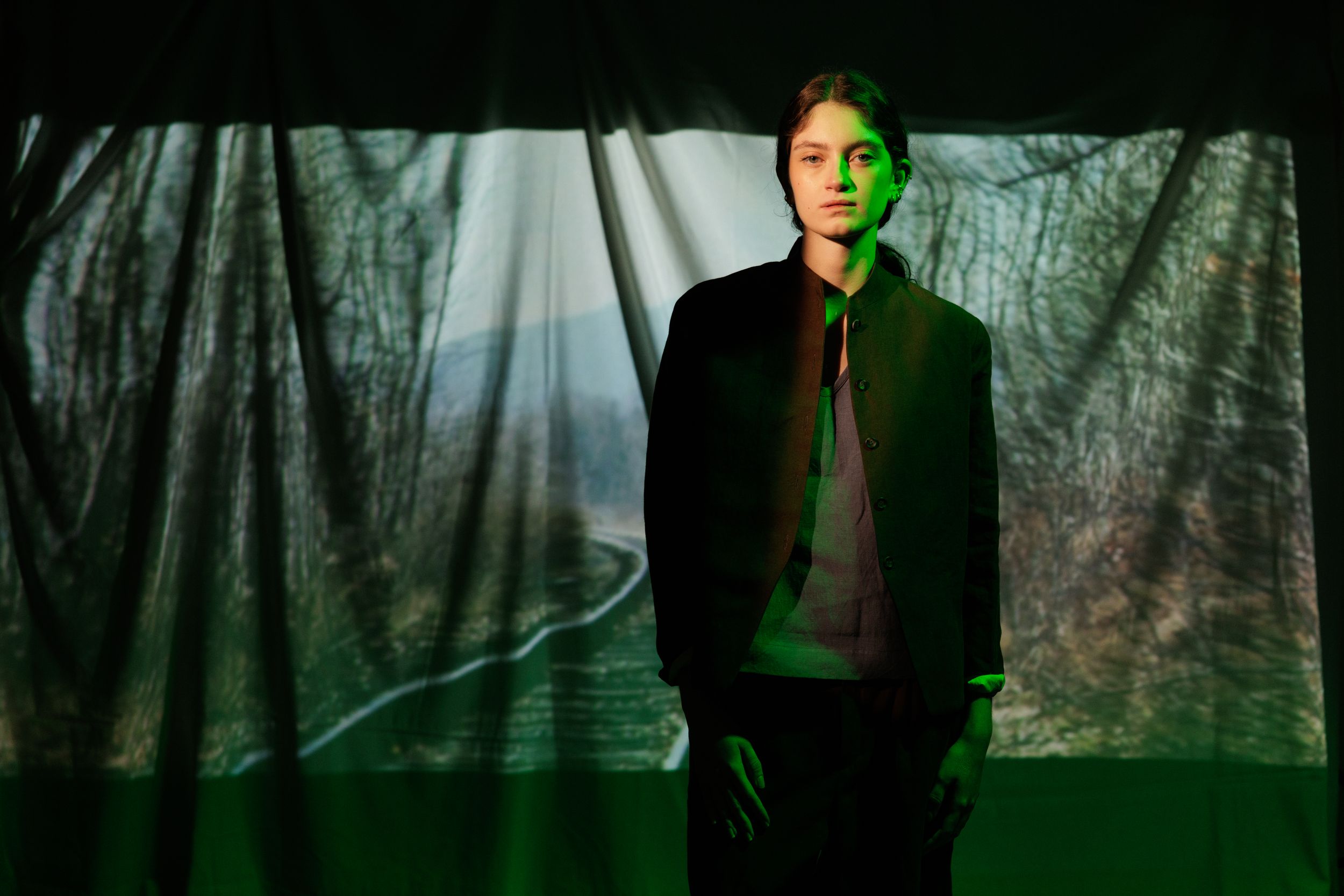 Figure illuminated by dramatic green lighting stands in front of dark textured backdrop with light beams shining through.