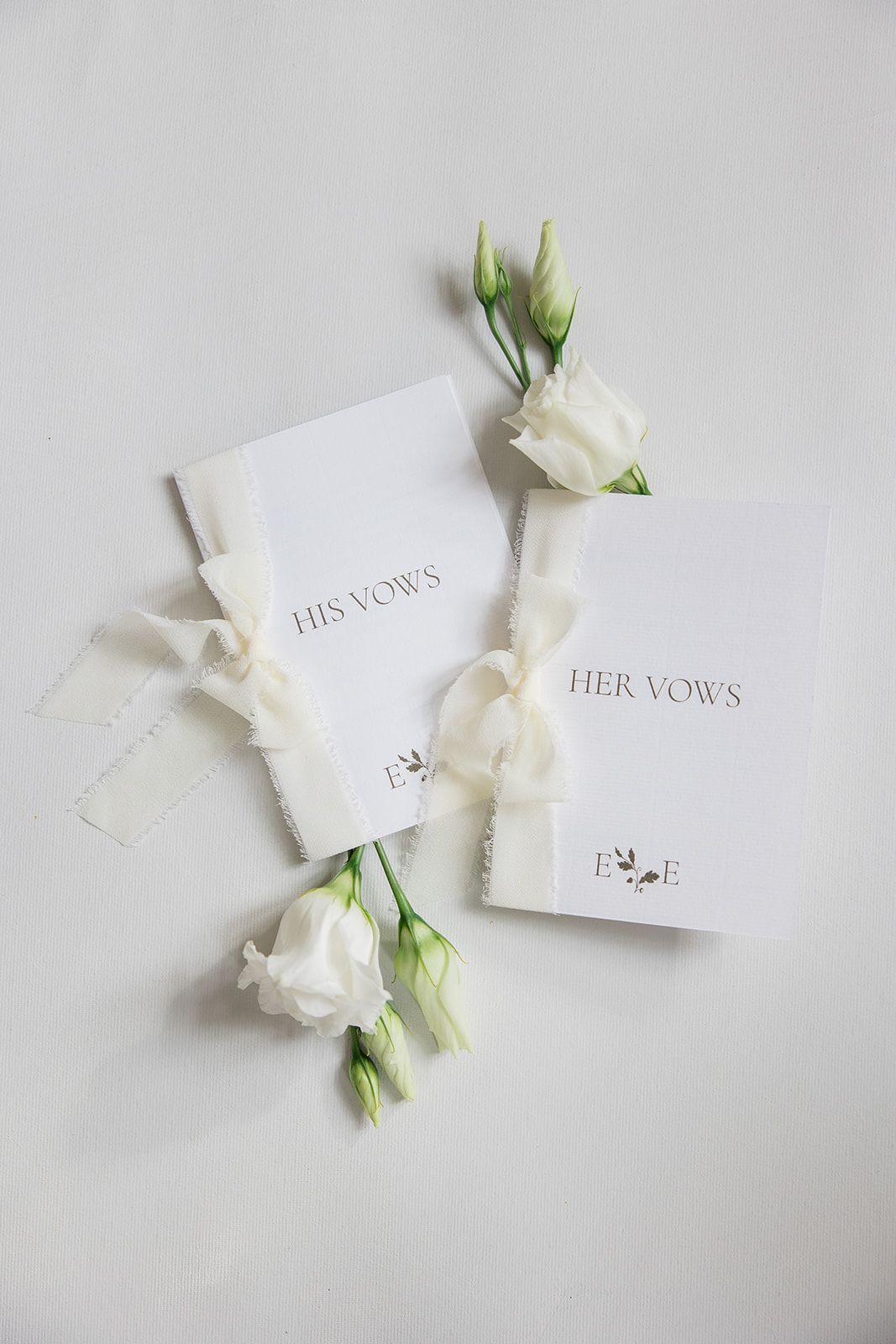Wedding vow booklets with 'His Vows' and 'Her Vows' text lie beside delicate white flowers on a light gray background.