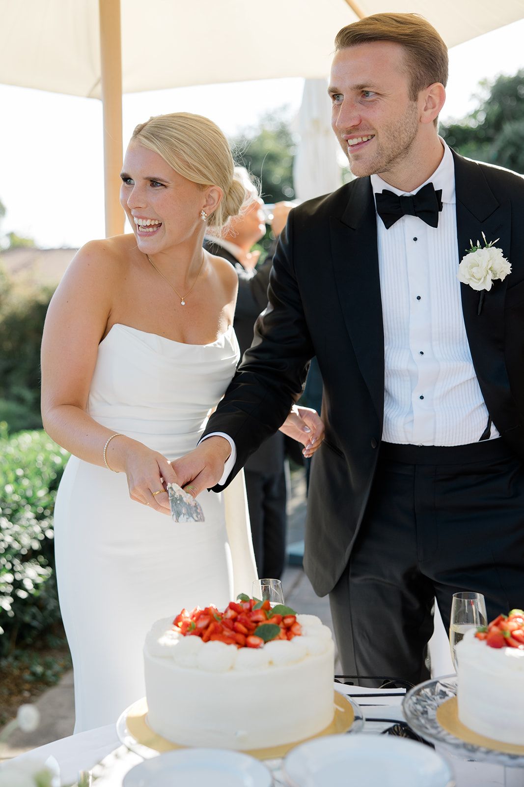 Newlyweds cut their elegant white wedding cake together during their outdoor reception celebration.